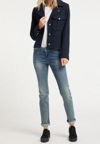Žena oblečená v námořnické sako na knoflíky, bílé košili, vybledlých modrých džínech s ohrnutými nohavicemi a černých platformových teniskách stojící na jednobarevném pozadí.