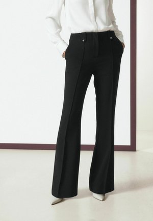 Persona che indossa pantaloni neri a vita alta e gamba larga con bottoni vicino alla vita, camicia bianca e scarpe bianche a punta, mani nelle tasche.