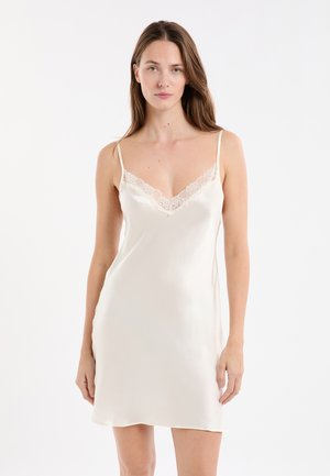 Donna che indossa un abito in satin color crema con pizzo sul collo, in piedi contro uno sfondo bianco.