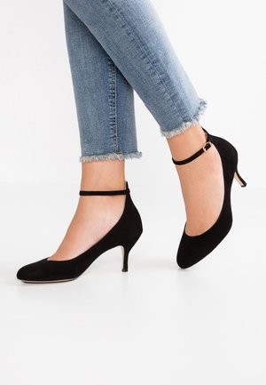 Černé semišové lodičky na vysokém podpatku mají špičatou špičku, kotníkový pásek se zlatými detaily a úzký podpatek, vystavené vedle džínsů s ohrnutými lemy.