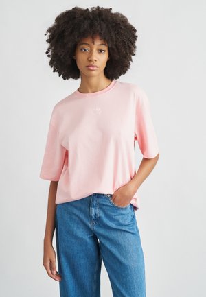 Svetloružové bavlnené tričko s krátkymi rukávmi a okrúhlym výstrihom, s malým bielym logom na hrudi. Kombinované s modrými rifľami.