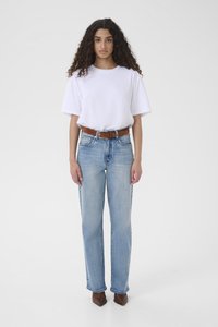 Keharõngastega naine, seljas valge t-särk, helesinised teksapüksid pruuni vööga ja teravnurksete pruunide kingadega, seisab ühevärvilise tausta ees.