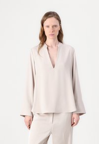 Vaalean beige pitkähihaisessa paidassa on avara pääntie, rento malli ja sileä pinta. Yhdistettynä samansävyisiin vaaleisiin housuihin.