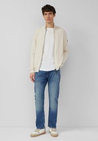 Lichtbeige bomberjack met een ritssluiting aan de voorkant, gecombineerd met een wit T-shirt en lichtblauwe jeans. Schoenen zijn van wit leer met beige accenten.