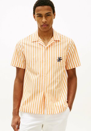 Młody mężczyzna w krótkim rękawie pomarańczowo-białej koszuli w pionowe paski z niebieskim wyszywanym logo na kieszeni na piersi.