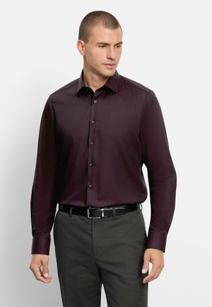 Mann trägt ein dunkelviolettes langärmeliges Hemd, das in graue Hose mit schwarzem Gürtel gesteckt ist, schaut leicht zur Seite.
