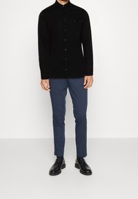 Camisa negra de manga larga con cuello puntiagudo, placket frontal y un pequeño logotipo. Combinada con pantalones azul marino de corte ajustado y zapatos negros.