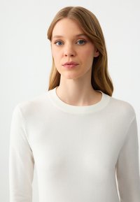 Maglione bianco con scollatura rotonda a coste, texture liscia, maniche lunghe e design aderente. Tessuto morbido con un leggero lucido.