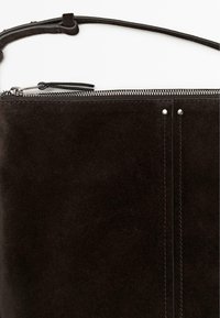 Dunkelbraune Umhängetasche aus Wildleder mit silbernem Reißverschluss, dünnem verstellbarem Riemen und vertikalen Nahtdetails mit Metallnieten.