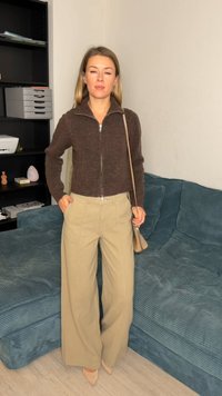 Pull brun à fermeture éclair, pantalon large beige clair, talons clairs et petit sac à bandoulière ; debout contre un canapé texturé couleur sarcelle.