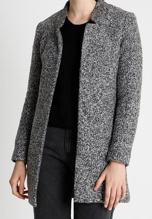 Femme portant un manteau long gris texturé par-dessus un pull noir et un pantalon foncé, debout devant un fond clair uni.