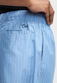 Χέρι ακουμπά στη μέση, φοράει μπλε και άσπρες κάθετες ριγέ σορτς με ελαστική μέση και πίσω τσέπη με κέντημα «Obey».