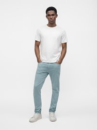 Jeune homme debout, les mains dans les poches, portant un t-shirt blanc uni, un pantalon bleu clair et des baskets blanches sur fond blanc.