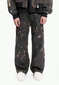 Person som har på seg løse mørke camouflasjekamuflerte cargobukser og hvite sko, står med hendene avslappet ved siden av seg foran en hvit bakgrunn.