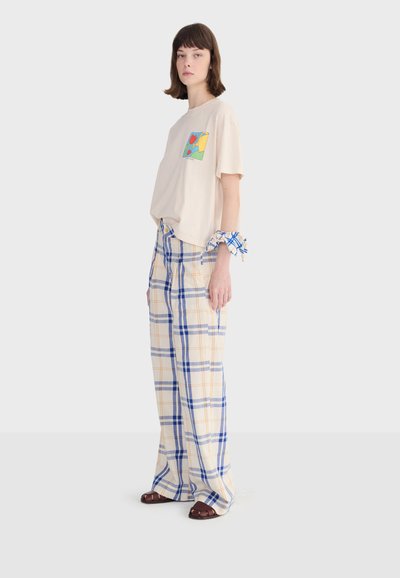 Femme portant un t-shirt graphique beige, un pantalon large à carreaux bleu et jaune, des sandales et un chouchou à carreaux au poignet, posant devant un fond blanc.