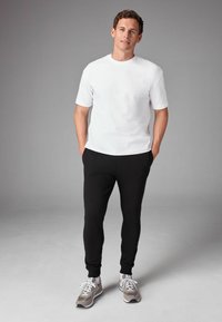 Weiße kurzärmlige T-Shirt, kombiniert mit schwarzen sportlichen Jogginghosen und grauen Sneakern. Das Outfit zeichnet sich durch eine lockere Passform und eine glatte Textur aus.