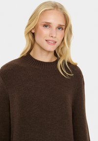 Maglione a coste marrone con scollatura rotonda, maniche lunghe e finitura texturizzata. Il tessuto appare morbido e caldo.