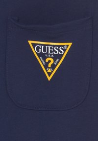 Navy niebieski materiał o teksturowanym wykończeniu z żółtym logo w kształcie trójkąta z napisem "GUESS U.S.A. WASHED JEANS" oraz wzorem znaku zapytania.