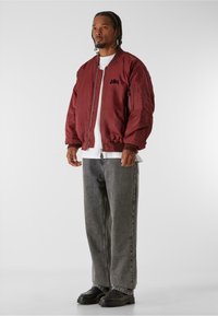 Bordový bombardér s předním zipem, žebrovanými manžetami a postranními kapsami. Nosený přes bílé tričko s šedými širokými kalhotami a černými botami.