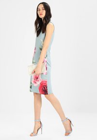 Blaues Bodycon-Kleid mit blauen, pinken und roten Blumenakzenten. Es hat einen ärmellosen Schnitt, eine knielange Länge und wird mit hellblauen High Heels und einer Clutch kombiniert.