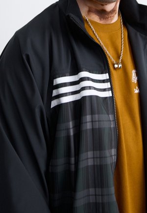 Personne portant une veste noire avec des rayures blanches sur une chemise moutarde et un collier chaîne argentée avec deux pendentifs.