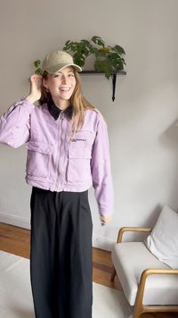 Casaco cropped roxo claro com colarinho preto, fecho na frente e dois bolsos, combinado com calças longas pretas e um boné bege.