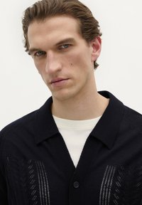 Cămașă albastră cu nasturi, cu guler, având model vertical din tricot și o textură moale. Purpă peste un tricou de culoare deschisă.