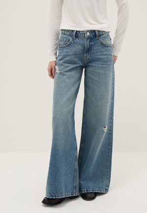 Persona vistiendo vaqueros de pierna ancha azul claro con pequeños desgarrones en el muslo y la rodilla, camiseta blanca de manga larga y zapatos negros.