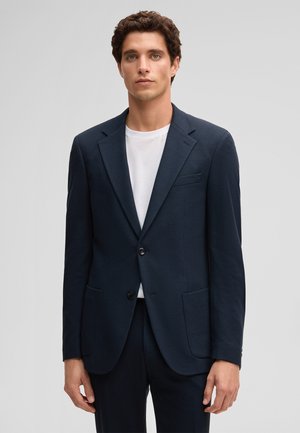 Młody mężczyzna w ciemnogranatowej marynarce na białej koszulce, stojący na tle jednolitego jasnoszarego tła.