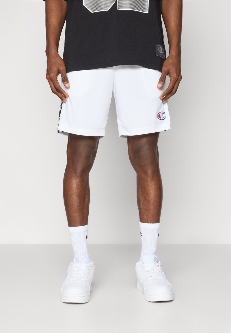 Calções atléticos brancos com um logótipo na perna esquerda, combinados com sapatilhas brancas e meias até ao tornozelo. Camiseta preta com uma grande estampa gráfica.