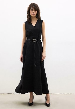 Vrouw met krullend bruin haar draagt een mouwloze zwarte maxi-jurk met een dunne riem en zwarte puntige hakken tegen een effen witte muur.