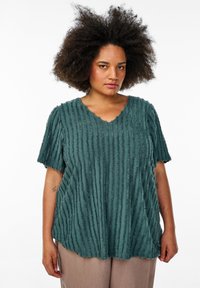 Top texturizado en color verde azulado con escote en V y mangas cortas. La tela presenta rayas verticales con una textura ligeramente deshilachada. Se lleva sobre pantalones claros.