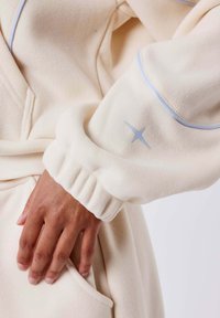 Cremfarbener Pullover mit gerippten Bündchen und hellblauen Paspeln. Verfügt über einen kleinen silbernen Sternelement an den Ärmeln und einen weichen, strukturierten Stoff.