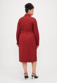 Vestiário vermelho, cintado, com colarinho, mangas longas e fendas laterais. Textura suave com um design até ao joelho. Combinado com sapatos de salto pretos.