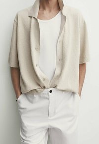 Beige gestructureerde kortmouwen cardigan met een kraag en knopen aan de voorkant over een witte aansluitende tanktop, gecombineerd met witte broek.