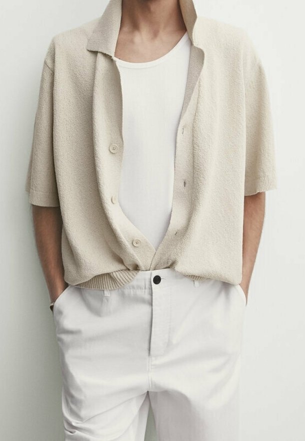 Beige gestructureerde kortmouwen cardigan met een kraag en knopen aan de voorkant over een witte aansluitende tanktop, gecombineerd met witte broek.