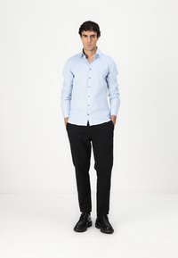Lichtblauw overhemd met lange mouwen en bruine knopen, gecombineerd met zwarte broek en zwarte schoenen, staande op een witte achtergrond.