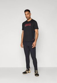 Vīrietis stāv pret vienkāršu fona attēlu, valkājot melnu ar grafiku apdrukātu T-kreklu, melnas bikses un melnas un bēšas krāsas augstpapēžu sporta apavus.