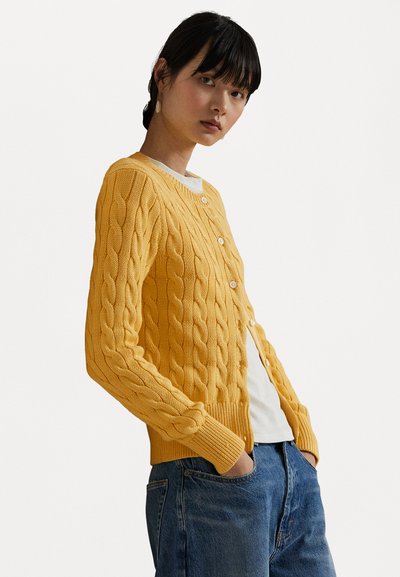 Gelber Kabelstrick-Cardigan mit geripptem Saum und Bündchen, ausgestattet mit fünf Knöpfen an der Vorderseite. Getragen über einem weißen Hemd und blauen Jeans.