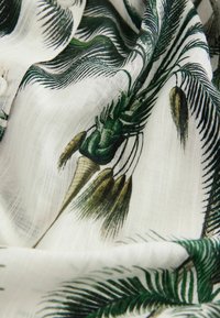Lichtgewicht stof met een groen palmbladpatroon op een witte achtergrond, met een gladde textuur en subtiele glans. Knopen zichtbaar.
