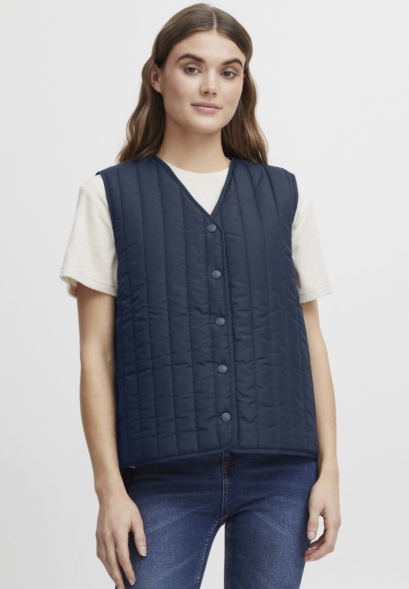 Marinblå quiltad väst med v-ringning, framknappar och sidofickor, bärs över en ljus t-shirt och visar en enkel och funktionell design.