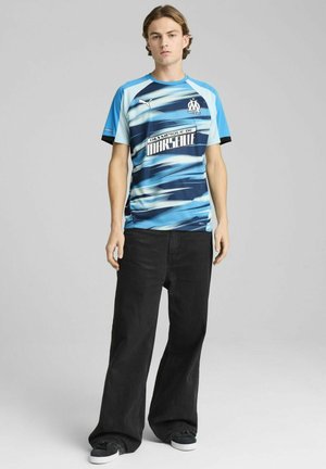 Blå og sort fodboldshirt med korte ærmer, bølgemønstre, logo og tekst på brystet, parret med sorte vidde bukser og sneakers.
