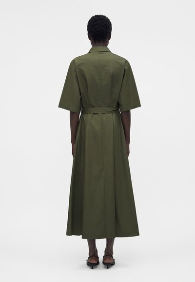 Γυναίκα με κοντά σγουρά μαλλιά, που φοράει ένα μίντι πράσινο-ελιά φόρεμα με ζώνη, μανίκια μέχρι τον αγκώνα και μαύρα σανδάλια με τακούνι, γυρισμένη πλάτη.