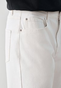 Gros plan sur la taille et la partie supérieure de la cuisse d'un jean en denim blanc avec poches visibles et passants de ceinture, porté avec un haut noir.