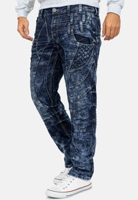 Jeans in denim con lavaggio blu sbiadito, texture strappata, tasche angolari, cuciture decorative e cuciture a contrasto. Abbinati a sneakers bianche.