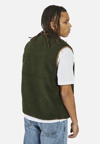 Colete de fleece verde com uma superfície texturada, decote redondo e design sem mangas. Usado sobre uma t-shirt branca e combinado com jeans azuis.