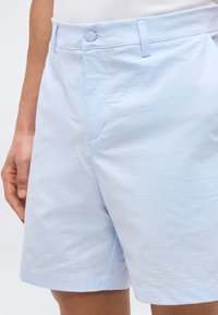 Σιέλ σορτς ψηλόμεσο με κουμπί και θηλιές για ζώνη, φορεμένο από ένα άτομο του οποίου φαίνεται το αριστερό χέρι, σε απλό φόντο.