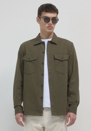 Mann trägt ein dunkelgrünes Hemd über einem weißen T-Shirt und hellbeige Hosen, mit dunklen Sonnenbrillen, steht vor einem schlichten Hintergrund.