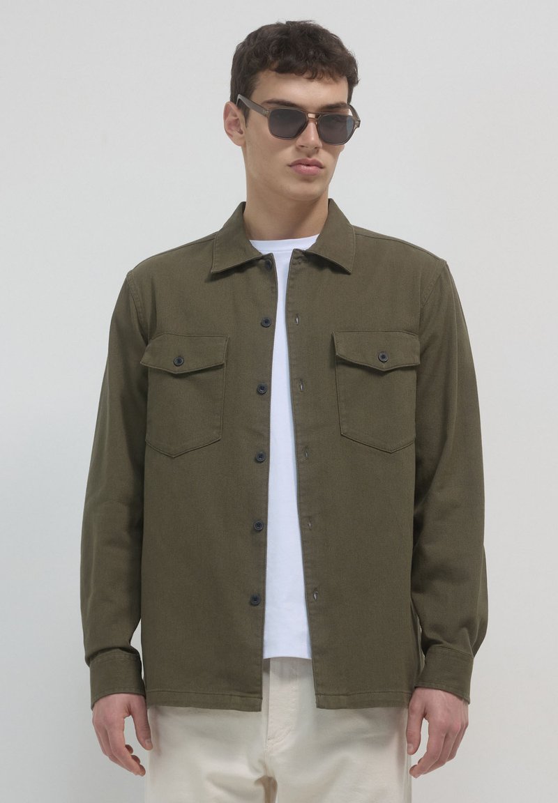 Uomo con camicia verde scuro a bottoni sopra una maglietta bianca e pantaloni beige chiaro, con occhiali da sole scuri, in piedi contro uno sfondo semplice.