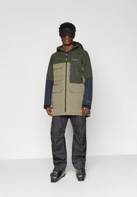 Vandeniui atspari striukė su kapišonu, alyvuogių žalios ir tamsiai mėlynos spalvos juostomis, kelios priekinės kišenės, juodos kelnės, dėvimos su juodais akiniais ir batais.